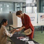 Duas mulheres interagem em um balcão de atendimento durante o dia de votação, em um ambiente eleitoral interno.