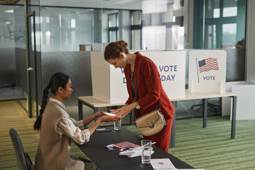 Duas mulheres interagem em um balcão de atendimento durante o dia de votação, em um ambiente eleitoral interno.